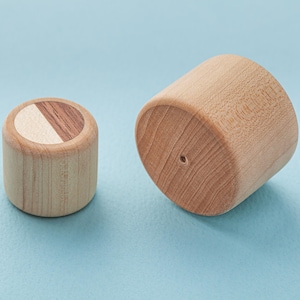 May include: Two wooden cylinders on a light blue surface. One is upright, with a top split between light and dark wood. The other is on its side, displaying the wood grain. The cylinders are made of wood.