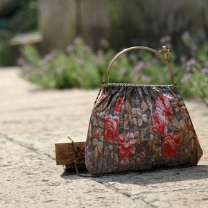 Può includere: Borsa a mano con stampa floreale, manico dorato e tracolla a catena. La borsa presenta un design a pieghe con un motivo di rose rosse e foglie su sfondo grigio. Una piccola scatola di legno è vicina.