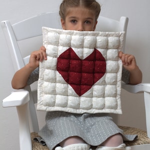 Può includere: Un cuscino trapuntato bianco con un design a cuore rosso. Il cuore è realizzato in tessuto rosso a pois bianchi. Il cuscino è tenuto da una persona seduta su una sedia a dondolo bianca.