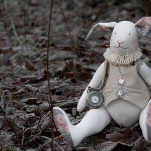 Può includere: Un coniglio di peluche bianco con un gilet marrone e un orologio da tasca. Il coniglio è seduto su un letto di foglie e ramoscelli marroni.