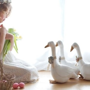 Può includere: Una bambina con un vestito bianco tiene un mazzo di fiori gialli mentre guarda tre statuine di oche bianche. Le oche sono in piedi su un pavimento di legno.