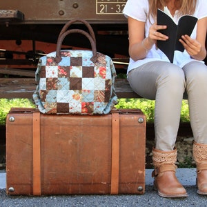 Può includere: Una donna è seduta su una panchina con una valigia di cuoio marrone e una borsa di tela patchwork. Sta leggendo un libro nero.