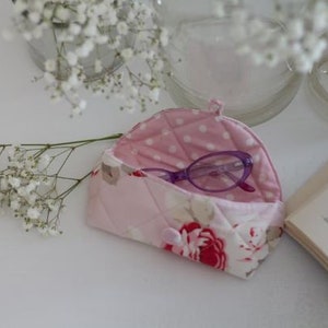 Può includere: Un astuccio per occhiali trapuntato rosa con un motivo floreale e un interno a pois. Occhiali viola riposano all'interno dell'astuccio. L'astuccio è aperto, rivelando gli occhiali. Fiori bianchi e un libro sullo sfondo.