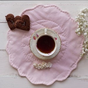 Può includere: Un tovaglietta rosa a forma di conchiglia con una tazza di tè e due biscotti a forma di cuore. La tazza da tè è bianca con motivi floreali e un bordo dorato. I biscotti sono marroni e hanno una forma di cuore.