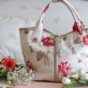 Può includere: Una borsa tote bianca con un motivo floreale con rose rosse. La borsa ha un manico in pelle marrone e una chiusura a zip.