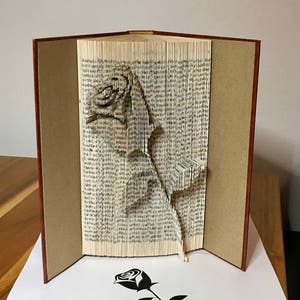 May include: A book art sculpture of a rose made by folding the pages of a book. The rose is brown and white and is standing on a wooden surface. The book is closed and has a brown cover. A black and white silhouette of a rose is printed on a white piece of paper below the book.
