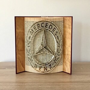 Pode incluir: Uma escultura de arte de livro do logotipo da Mercedes-Benz. O logotipo é feito de páginas dobradas de um livro e é exibido em uma superfície de madeira.