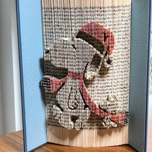May include: A book art sculpture of a dog wearing a red hat and scarf. The sculpture is made from the pages of a book that have been folded and shaped to create the image. The sculpture is displayed on a blue background with white polka dots.