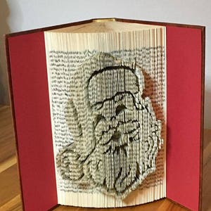 Peut inclure: Un livre dont les pages sont pliées en une sculpture 3D d'un visage humain. Le livre a des couvertures rouges et la sculpture est faite de papier blanc.