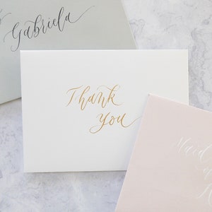 May include: Three envelopes with calligraphy on them. The top envelope is light grey with black calligraphy that says "to Gabriela". The middle envelope is white with gold calligraphy that says "Thank you". The bottom envelope is light pink with white calligraphy that says "Maid of Honor".