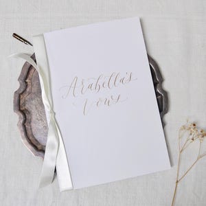 May include: A white booklet with a gold ribbon tied around it. The cover has the words "Arabella's Vows" written in elegant calligraphy.