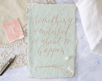 May include: A light green card with a handwritten message in gold lettering that reads "Something wonderful is about to happen". The card is decorated with dried white flowers.
