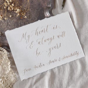 May include: A white paper with a deckled edge displays brown calligraphy. The quote reads "My heart is, & always will be yours, Jane Austen - Sense & Sensibility." The paper is on a silver tray, with a pen and dried flowers.