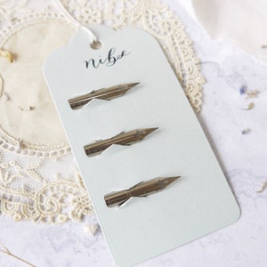 May include: Three silver calligraphy nibs on a white tag with the word "nibe" written in black calligraphy.