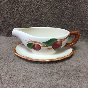 May include: A white ceramic gravy boat with a matching saucer. The boat has a brown handle and is decorated with a branch design featuring red plums and green leaves. The rim of the saucer is brown.