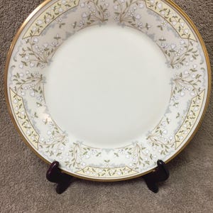 May include: A white porcelain plate with a gold rim and a floral design. The plate features a cream-colored border with a delicate pattern of white flowers and green leaves. The plate is displayed on a dark brown stand.