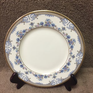 May include: A white porcelain plate with a gold rim and a floral design in blue and pink. The plate is supported by a dark brown wooden stand, showcasing a classic and elegant aesthetic.