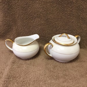May include: A white porcelain creamer and sugar bowl set. The creamer has a curved spout and handle. The sugar bowl has a lid and two handles. Both pieces have gold trim and a thin gold band around the middle.