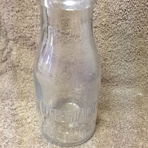 May include: A clear glass milk bottle with a rounded body and a narrow neck. The bottle has the embossed text "HEBERHART" near the base. The bottle is empty and sits on a textured surface.