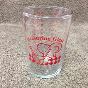 May include: Clear measuring glass with red markings and a red graphic of a chef's hat, whisk, and spoon. The glass has measurements in cups, tablespoons, and teaspoons. The text "Measuring Glass" is printed in red.