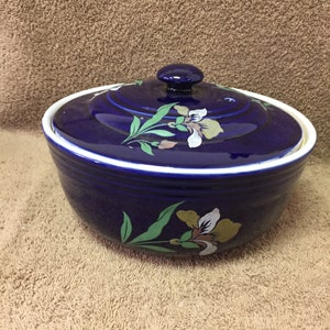 May include: A cobalt blue ceramic casserole dish with a lid. The dish features a white rim and is decorated with floral designs in green, white, and gold. The lid has a small, round knob.
