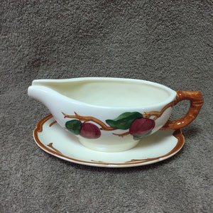 May include: Cream-colored ceramic gravy boat with a matching saucer. The boat features a brown handle and is decorated with a branch design with red and green fruit. A classic serving piece for the dining table.