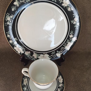 May include: A black and white floral patterned tea set. The set includes a large plate, a teacup, and a saucer. The plate and saucer have a black border with white floral designs and gold trim. The teacup is white with a gold trim.