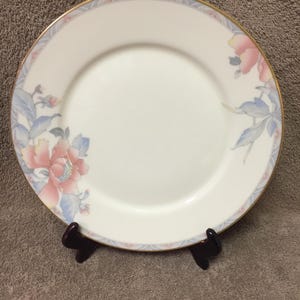 May include: White porcelain plate with a gold rim and a floral design featuring pink and blue flowers. The plate has a light blue and pink border. The plate is displayed on a dark brown stand.
