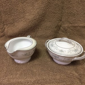 Puede incluir: Un juego de crema y azucarero con tapa de porcelana blanca. Ambas piezas presentan un delicado estampado floral en azul claro y gris, con ribete plateado. La cremera tiene un pico y un asa, mientras que el azucarero tiene tapa y asas.