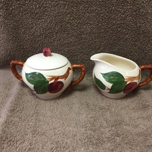 May include: A cream-colored ceramic sugar bowl with a lid and a creamer. Both pieces feature a hand-painted design of red apples and green leaves. The handles are brown. The sugar bowl has a red knob on the lid.