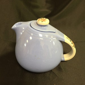 May include: A light blue ceramic teapot with a rounded body, a curved spout, and a handle with floral accents. The lid features a floral design on the knob. The teapot is designed for serving tea.