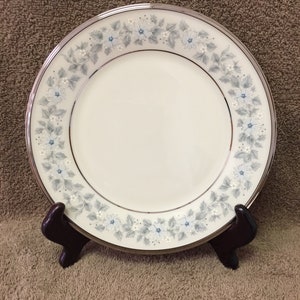 May include: A white ceramic plate with a silver rim and a floral pattern in light blue and gray. The plate is displayed on a dark brown stand, showcasing its decorative design. The plate's diameter is approximately 10 inches.