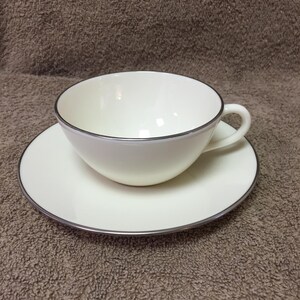 May include: A white teacup and saucer set with a silver trim. The teacup has a curved handle and sits on a matching saucer. The set is made of porcelain and is suitable for serving tea or coffee.