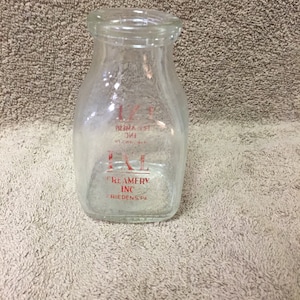 May include: A clear glass milk bottle with a square base and a narrow neck. The bottle has red text that reads "1/2 PT" and "I.L. CREAMERY INC. FRIEDEN'S, PA."