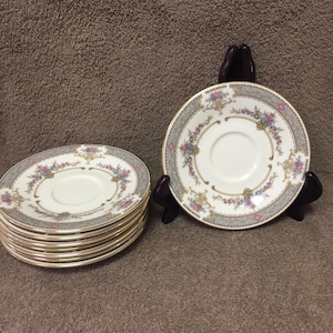 May include: A set of vintage porcelain saucers. Each saucer features a white center with a floral design in pink, purple, and blue, framed by a gray border and gold trim. The saucers are stacked and displayed on a brown surface.