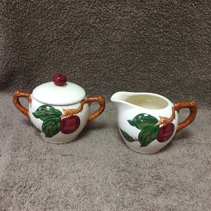 May include: A white ceramic sugar bowl with a lid and a matching creamer. Both pieces feature a design of red apples and green leaves. The handles are brown. The sugar bowl lid has a red knob.
