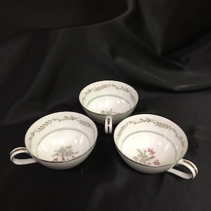 May include: Three white porcelain teacups with handles. Each cup features a floral design with pink and green accents, a thin blue line, and a gold rim. The cups are arranged on a black surface.