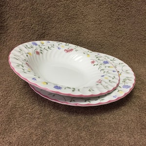 May include: Two white oval serving dishes with a floral design. The dishes have a pink trim and a pattern of flowers in pink, blue, and yellow. The dishes are stacked on top of each other.
