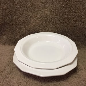 May include: Two white octagonal ceramic dishes, one a shallow bowl and the other a plate, are stacked on a brown surface. The dishes have a simple, clean design, suitable for serving food. The edges of the dishes are slightly raised.
