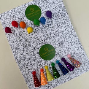 May include: A selection of rainbow-coloured party decorations, including balloon-shaped ornaments and glittery cone-shaped party hats. The items are arranged on a white background with black polka dots.