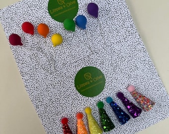 May include: A collection of colorful party decorations, including rainbow-colored balloon-shaped ornaments and glittery cone-shaped party hats. The items are arranged on a white background with black polka dots.