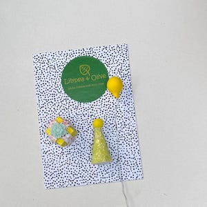 May include: A playful composition showing a yellow balloon on a wire, a party hat, and a gift box with a floral detail. These items are arranged on a card with a black and white polka dot design. The card displays a green circle with the text "Lilypea & Olive".