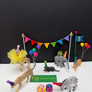 May include: A birthday party scene with toy animals, such as a giraffe, cheetah, elephant, tiger, and zebra, wearing party hats. The scene includes colourful bunting, balloons, and gift boxes. The banner says "Lilypea + Olive".