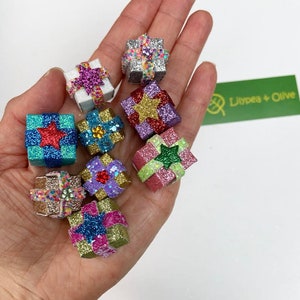 May include: A selection of small, glittery gift boxes in shades like silver, blue, and purple. Each box is decorated with a star or bow, and held in a hand. The Lilypea + Olive logo is visible in the background.
