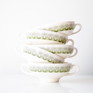 Peut inclure: Une pile de cinq tasses à soupe crème, ornées d'un motif géométrique vert et noir sur le bord. Chaque tasse est dotée d'une anse et est empilée, créant un agencement visuellement attrayant.