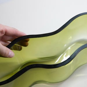 May include: An olive-green glass bowl with a black rim and a wavy design. The bowl's shape and colour create a modern aesthetic. The bowl is held by a hand.