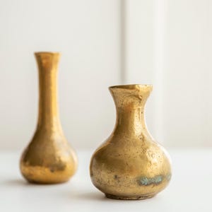 May include: Two brass vases with a metallic gold finish and a weathered, textured surface. One vase is tall and slender with a narrow neck, while the other is shorter and bulbous with a wider base and a small opening at the top.
