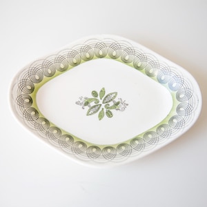Peut inclure: Plat à servir en céramique blanche, de forme losange aux bords arrondis. Le bord présente un motif répétitif de spirales noires et grises avec une bordure verte. Le centre a un motif floral vert et gris.