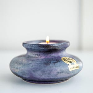 May include: A low, wide ceramic candle holder with a mottled purple and blue glaze. A small lit candle sits in the center opening. A gold-colored circular sticker on the side features the text Ruscha Art and the words echt Handarbeit.