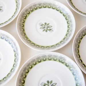 Peut inclure: Vue aérienne de plusieurs soucoupes blanches avec une bordure décorative verte et noire. Chaque soucoupe présente un motif floral central vert et gris. Les soucoupes sont disposées sur une surface blanche.